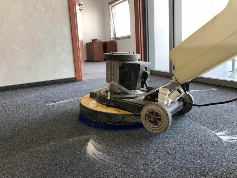 Maszyna z niebieską szczotką czyści szary dywan w biurze, pozostawiając za sobą mokre ślady.