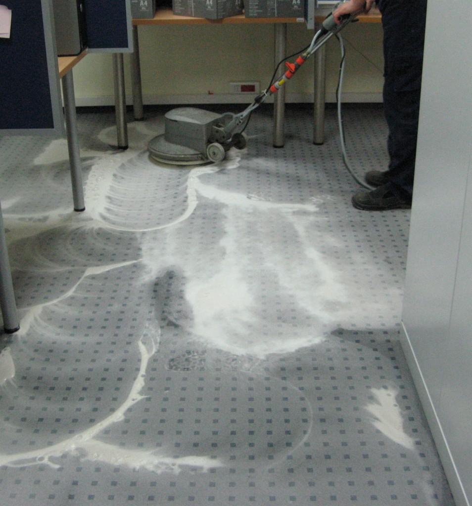 Szorowarka do podłóg w trakcie czyszczenia szarej wykładziny dywanowej w biurze, widoczne ślady mokrej piany i częściowo oczyszczony fragment podłogi.