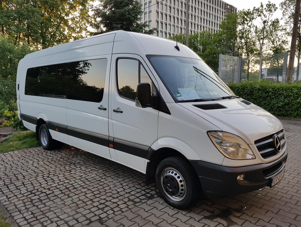 Biały, długi bus marki Mercedes z przyciemnianymi szybami, zaparkowany na kostce brukowej przed budynkiem i zielenią.