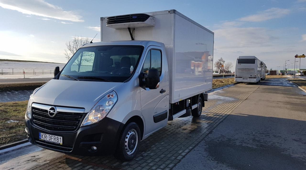 Srebrny Opel Movano z chłodnią na parkingu z widocznym autobusem w tle, tablica rejestracyjna KR 3FR81.