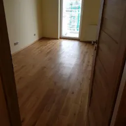 Pusty pokój z nową, ułożoną podłogą z paneli drewnianych, widok na okno z budową w tle i częściowo otwarte drzwi z drewna po prawej stronie.