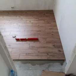 Układanie płytek imitujących drewno w pomieszczeniu, widoczny poziomica i fragment podłogi przed położeniem płytek, butelka wody i miarka w rogu.