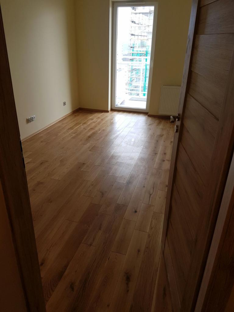 Pusty pokój z nową, ułożoną podłogą z paneli drewnianych, widok na okno z budową w tle i częściowo otwarte drzwi z drewna po prawej stronie.