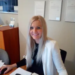 Uśmiechnięta blondynka w biurze, w tle dokumenty w ramkach, kalkulator i plany na biurku. Jasna marynarka i profesjonalny wygląd.