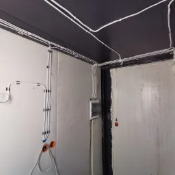 Wnętrze kontenera mieszkalnego z widocznymi, nowo poprowadzonymi białymi przewodami elektrycznymi, przymocowanymi do ściany i sufitu za pomocą czarnych uchwytów, oraz pomarańczowymi puszkami...