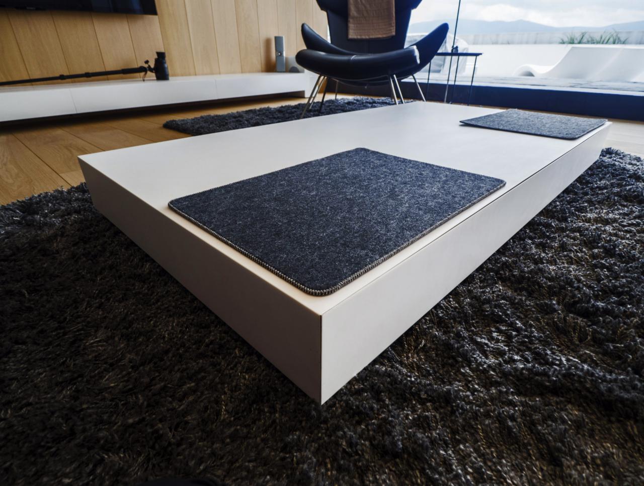 Nowoczesny, biały stolik kawowy z ciemnoszarymi podkładkami na grubym, ciemnym dywanie, w tle widoczne minimalistyczne meble i panorama za oknem.
