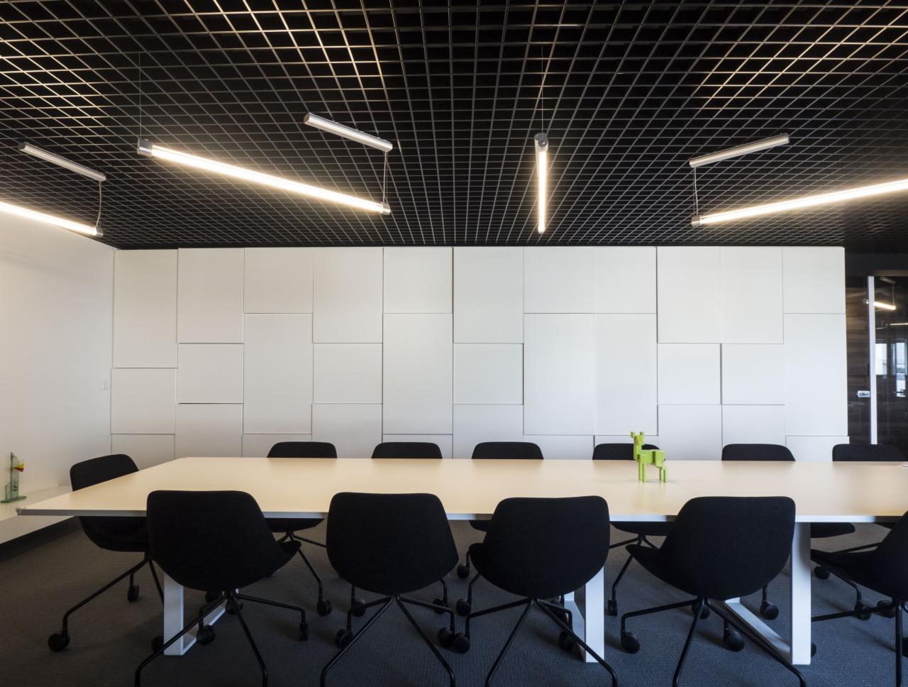 Nowoczesna sala konferencyjna w Świętej Katarzynie z długim stołem, czarnymi krzesłami i białą ścianą z paneli. Sufit rastrowy z podwieszonymi lampami liniowymi.