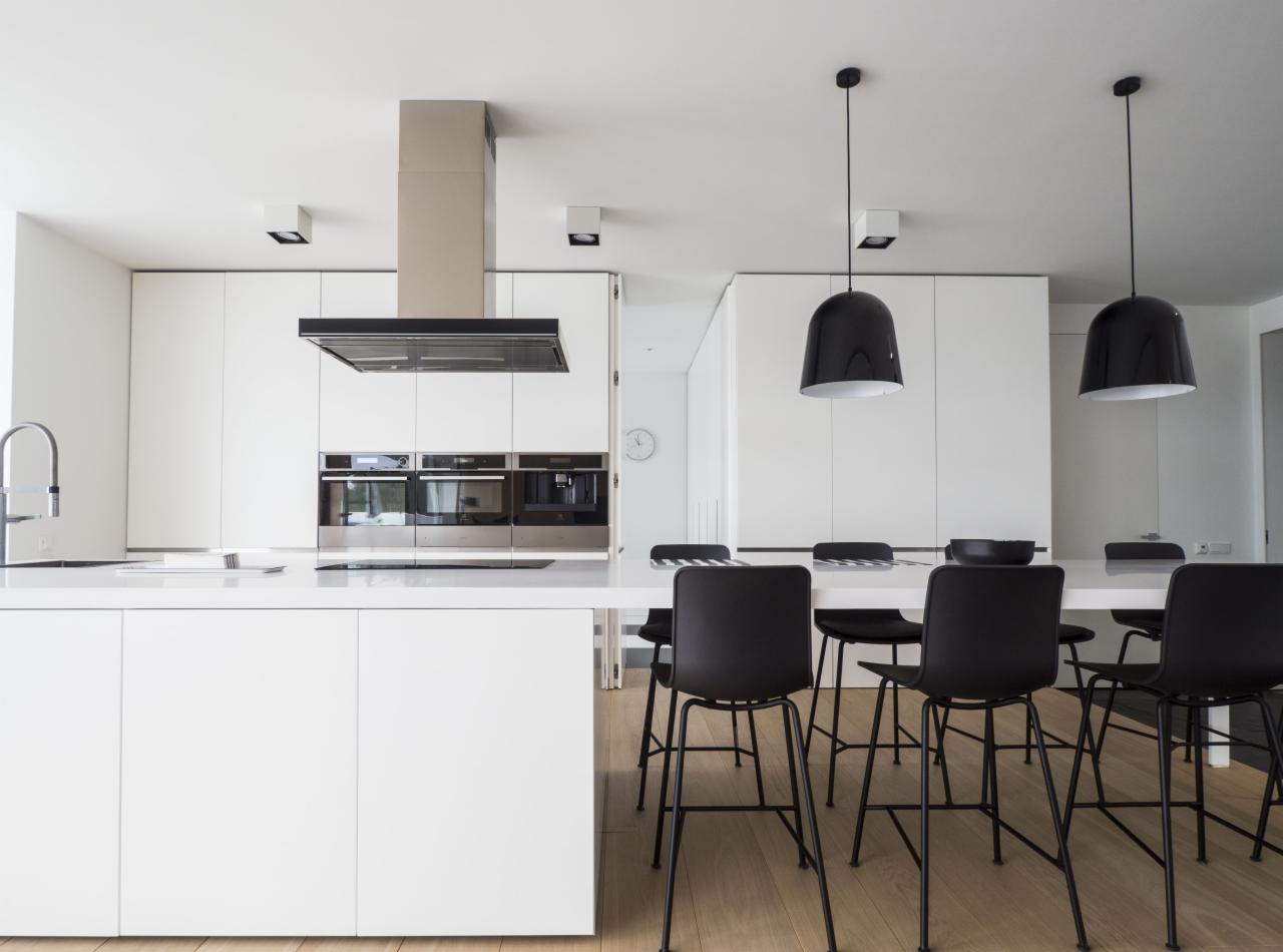 Minimalistyczna, nowoczesna kuchnia z białymi szafkami bez uchwytów, wyspą z blatem roboczym i czarnymi krzesłami barowymi, oświetlona trzema czarnymi lampami wiszącymi w kształcie kopuł.