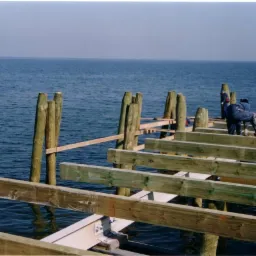 Konstrukcja pomostu na wodzie w trakcie budowy, widoczne drewniane belki i pale, pracownik w kombinezonie.