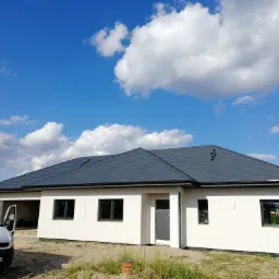 Nowoczesny dom jednorodzinny z grafitowym dachem z dachówki ceramicznej i czarnymi rynnami, widok z boku z samochodem dostawczym na podjeździe, błękitne niebo z obłokami w tle.