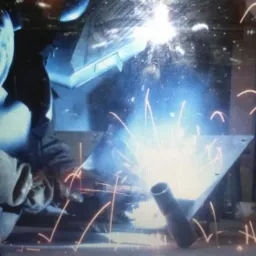 Spawacz w flanelowej koszuli łączy metalowe elementy, iskrząc jasnym światłem w ciemnym warsztacie.