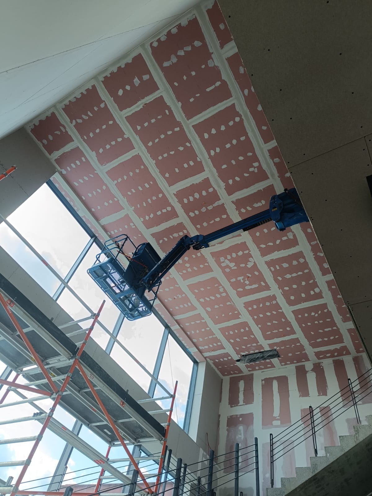 Montaż płyt gipsowo-kartonowych na skośnym suficie w nowoczesnym wnętrzu, widoczny podnośnik i rusztowania, duże okno z widokiem na niebo.