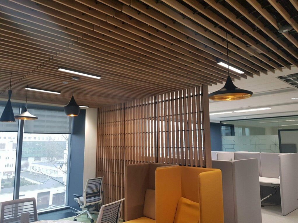 Nowoczesne biuro z drewnianym ażurowym sufitem i ścianką działową. Widoczne lampy wiszące i stanowiska pracy.