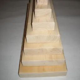 Stos ułożonych bloków z jasnego drewna o różnych wymiarach, widoczne słoje, ustawione piramidalnie na białym tle.