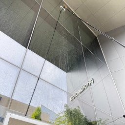 VERSA SYSTEM Przemysław Piechocki - Zewnętrzne mycie szklanej elewacji budynku z użyciem specjalistycznego sprzętu teleskopowego i wody. Widoczne refleksy otoczenia na czyszczonej powierzchni.