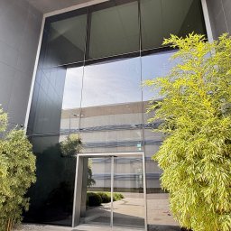 VERSA SYSTEM Przemysław Piechocki - Szklana fasada biurowca z odbiciem otoczenia i roślinami. Widoczne czyste szyby i wejście. Minimalistyczny design budynku.
