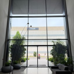 VERSA SYSTEM Przemysław Piechocki - Wnętrze biurowca z wysokimi oknami, widok na zieleń i budynek Empik. Szara sofa i lampa łukowa w holu. Duże okna zapewniają naturalne światło.