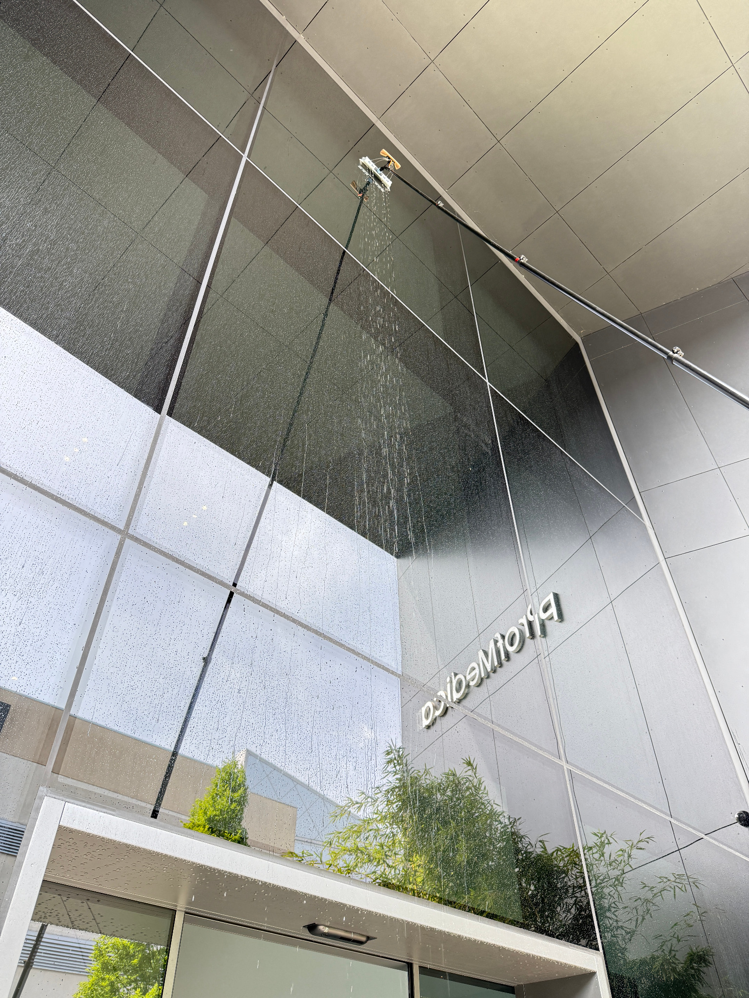 Zewnętrzne mycie szklanej elewacji budynku z użyciem specjalistycznego sprzętu teleskopowego i wody. Widoczne refleksy otoczenia na czyszczonej powierzchni.