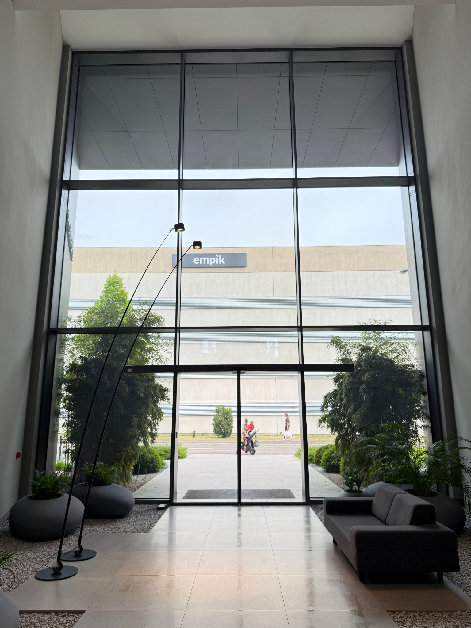Wnętrze biurowca z wysokimi oknami, widok na zieleń i budynek Empik. Szara sofa i lampa łukowa w holu. Duże okna zapewniają naturalne światło.