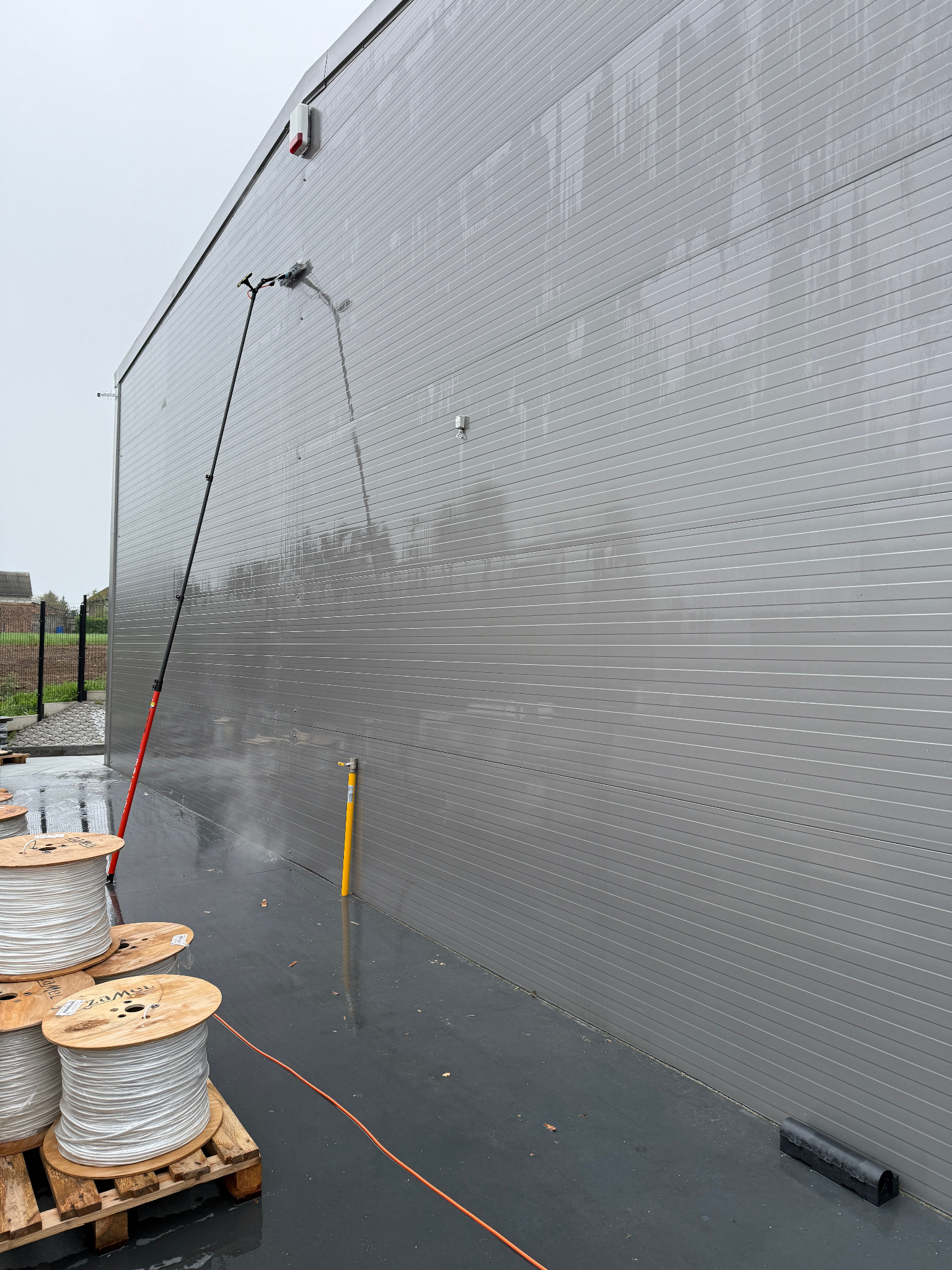 Mycie szarej elewacji przemysłowej za pomocą lancy teleskopowej. Na pierwszym planie drewniane szpule z białym kablem, mokra posadzka i elementy krajobrazu w tle.