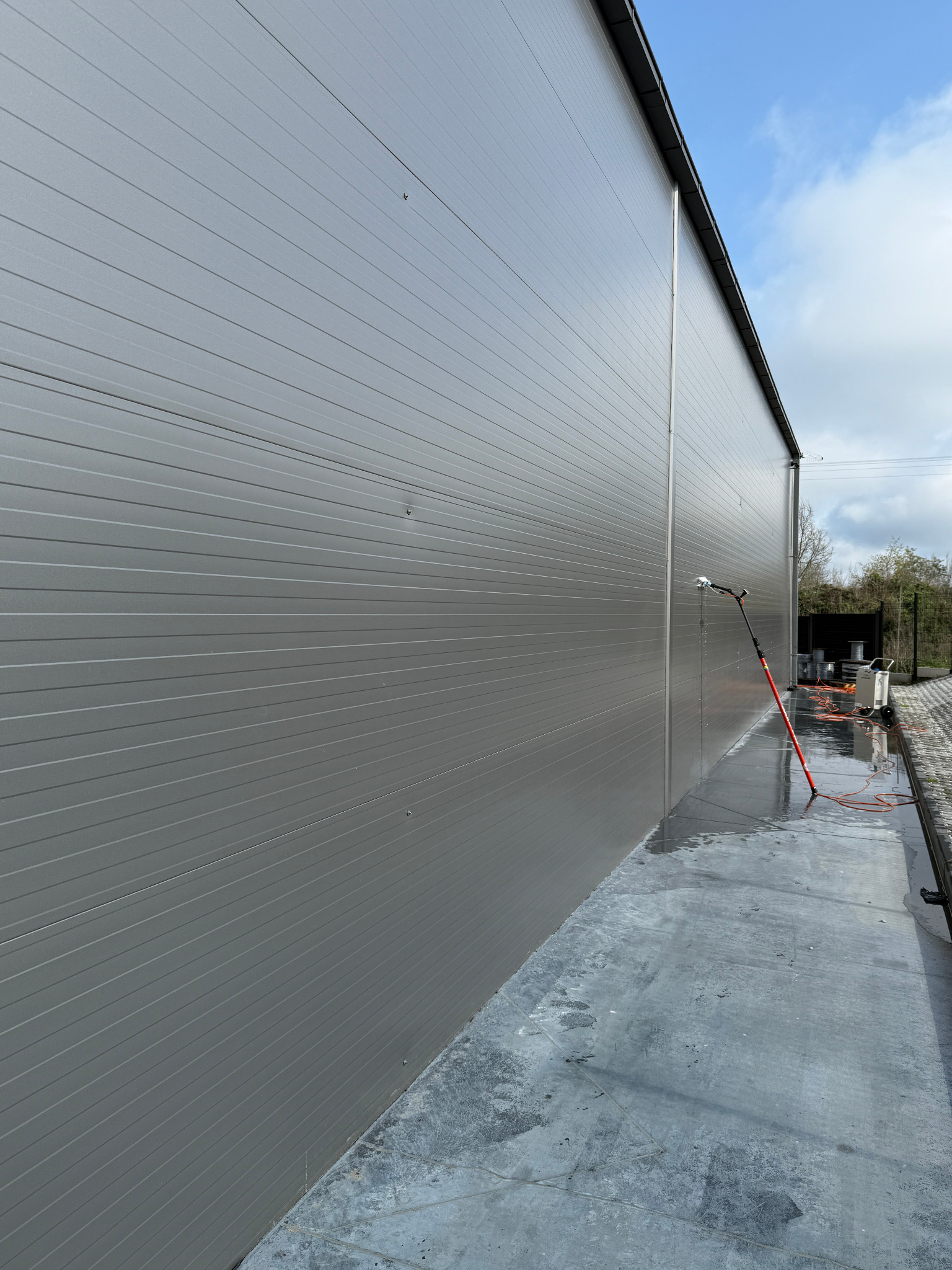 Szczegółowe ujęcie mycia szarej elewacji budynku przemysłowego za pomocą lancy teleskopowej, z widocznymi smugami wody i częściowo umytą powierzchnią.