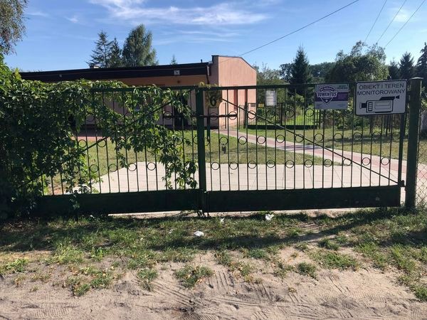 Zielona brama wjazdowa z okrągłymi zdobieniami, częściowo porośnięta bluszczem, z tabliczkami informacyjnymi o monitoringu i firmie, na tle budynku i drzew.