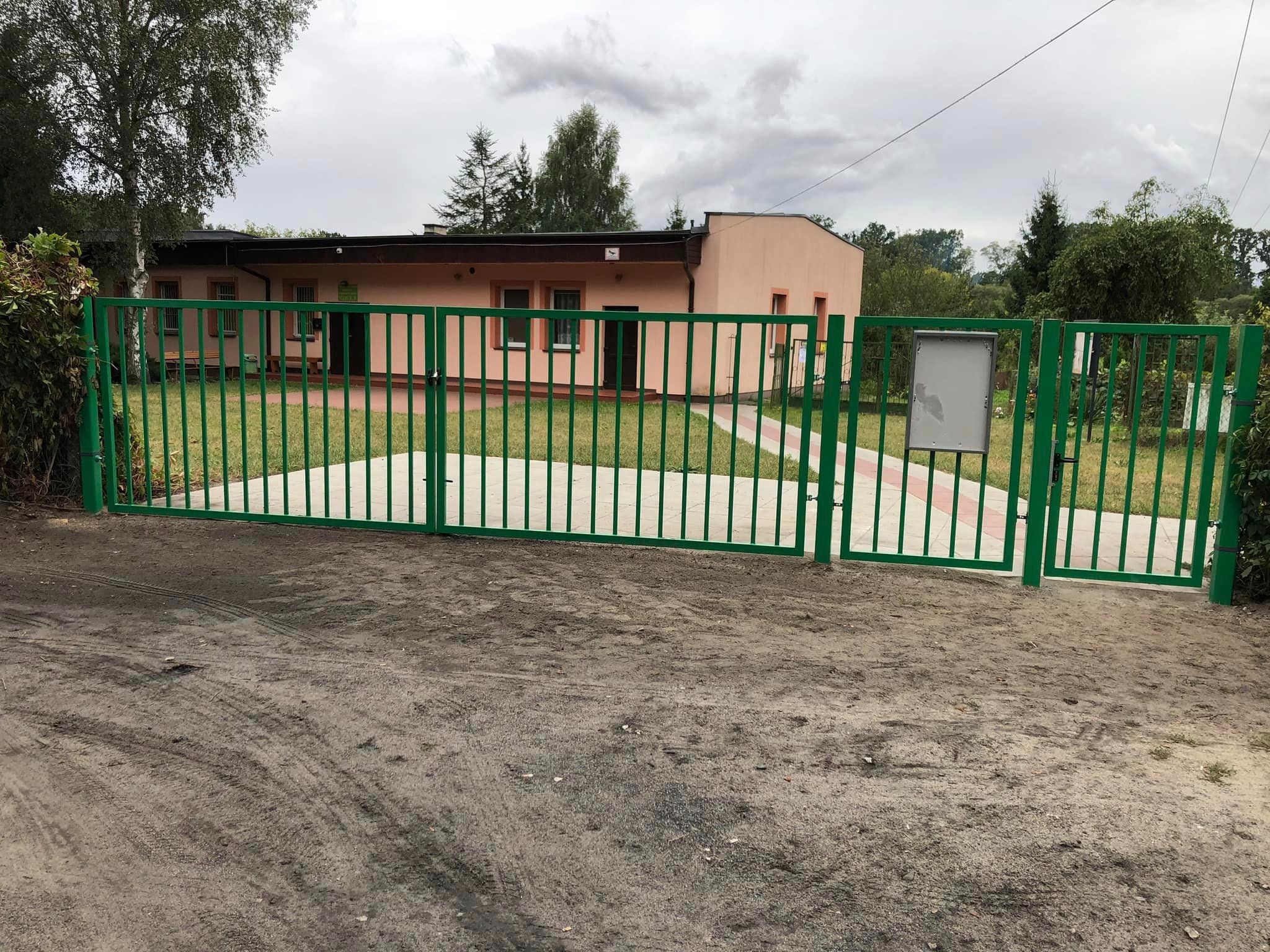 Zielona brama wjazdowa z pionowymi prętami, prowadząca do budynku z czerwono-białym chodnikiem, widok na pochmurne niebo.