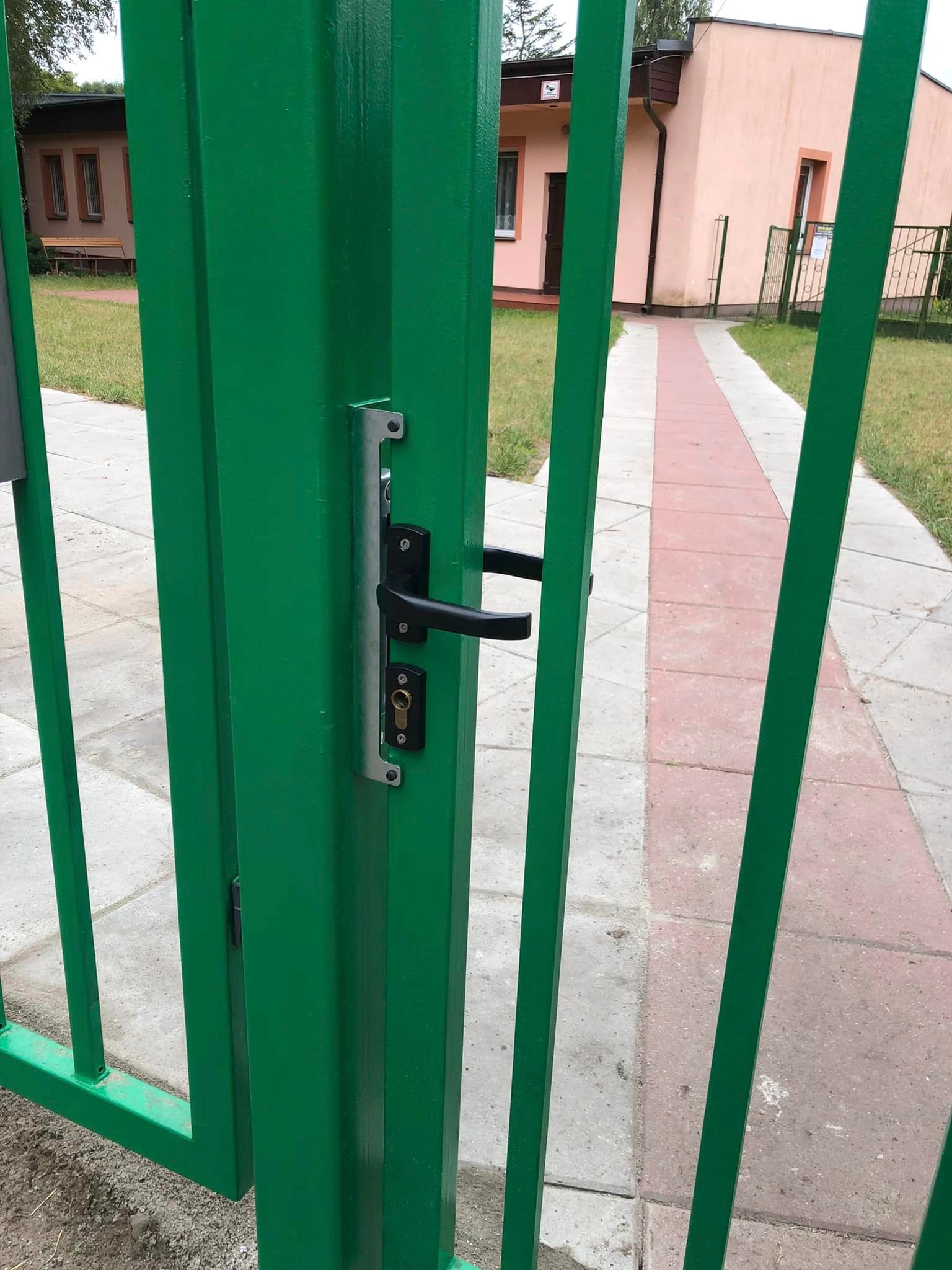 Zielona brama wjazdowa z czarną klamką i zamkiem, widok z bliska na mechanizm otwierania na tle budynku i chodnika.