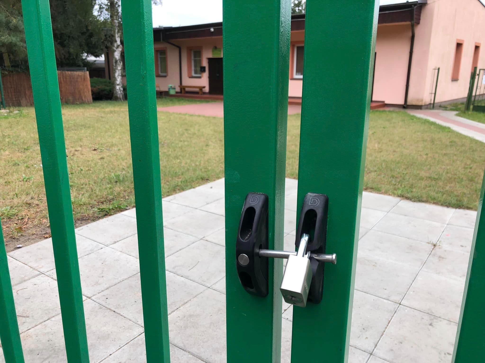 Zielona brama wjazdowa zabezpieczona srebrną kłódką, widok na trawnik i budynek w tle.