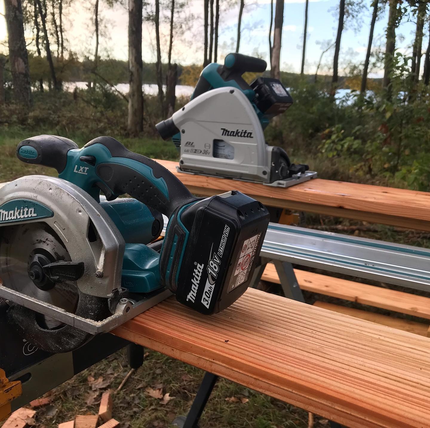 Dwie piły tarczowe akumulatorowe Makita na drewnianym blacie roboczym, w tle widok na jezioro i drzewa.