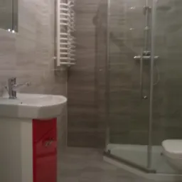 Nowoczesna łazienka z szarymi płytkami imitującymi drewno, białą umywalką z szafką w kolorze czerwonym, grzejnikiem drabinkowym i kabiną prysznicową typu walk-in.