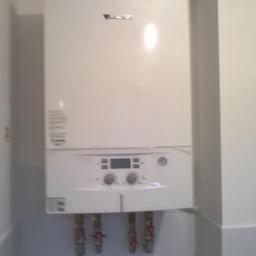 Usługi remontowo instalacyjne - Biały piec gazowy marki Vaillant, zamontowany na białej ścianie z widocznymi rurami i zaworami, z białym kominem odprowadzającym spaliny zakończonym niebieskim elementem.