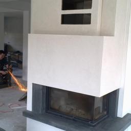 Usługi remontowo instalacyjne - Wnętrze w trakcie wykańczania z nowoczesnym kominkiem obudowanym białym tynkiem strukturalnym i ciemnym kamieniem, w tle pracownik używa szlifierki kątowej z iskrami.