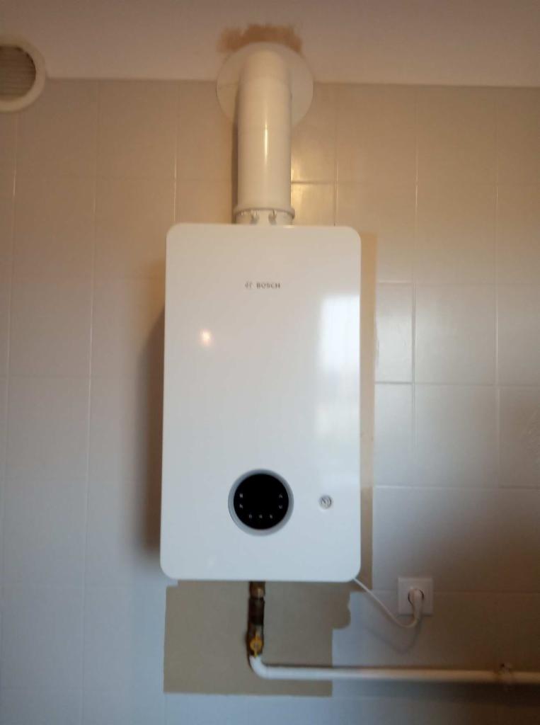 Biały piec gazowy marki Bosch zamontowany na ścianie z jasnymi kwadratowymi płytkami, widoczne rury doprowadzające i odprowadzające gaz oraz przewód zasilający.