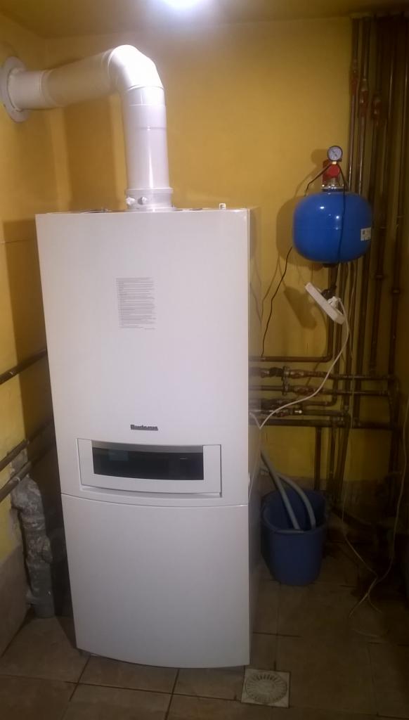Biały piec gazowy marki Buderus z podłączonymi rurami odprowadzającymi spaliny, niebieski zbiornik wyrównawczy ciśnienia oraz widoczne rury instalacji grzewczej na tle żółtej ściany.