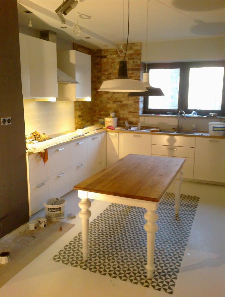 Wnętrze kuchni w trakcie prac wykończeniowych: białe szafki, ceglana ściana, wiszące lampy w stylu industrialnym, stół z drewnianym blatem na tle wzorzystej podłogi, widoczne puszki z farbą.