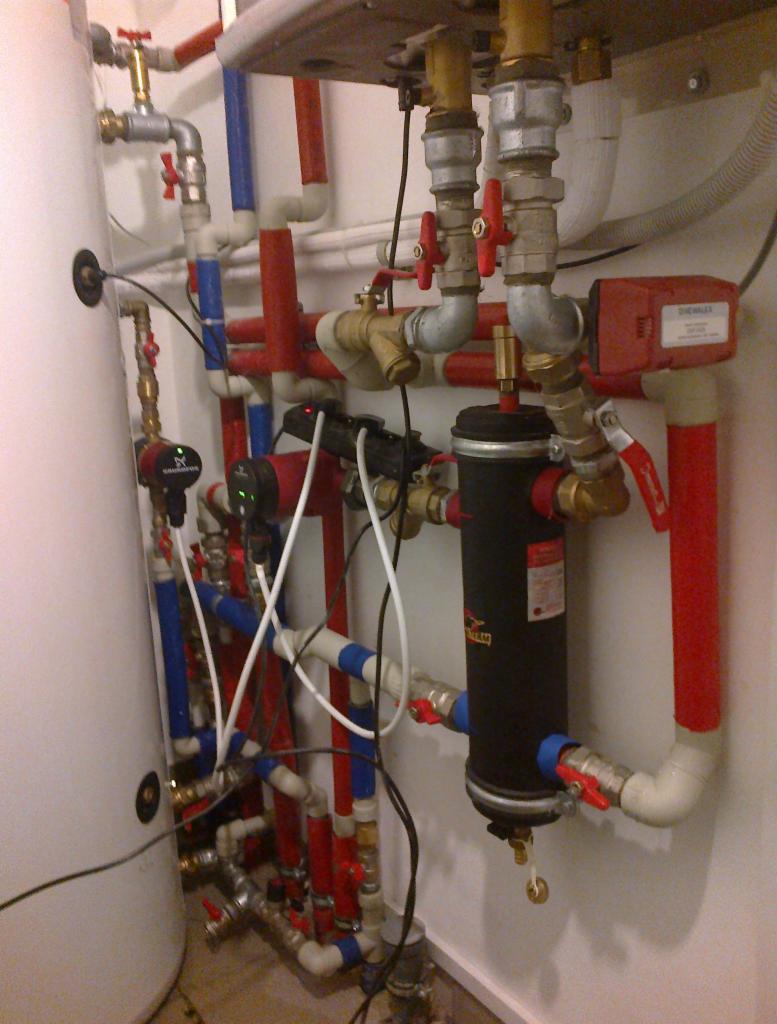 Złożona instalacja grzewcza z rurami w kolorze czerwonym, niebieskim i białym, licznymi zaworami i filtrem cylindrycznym, zamontowana na białej ścianie.
