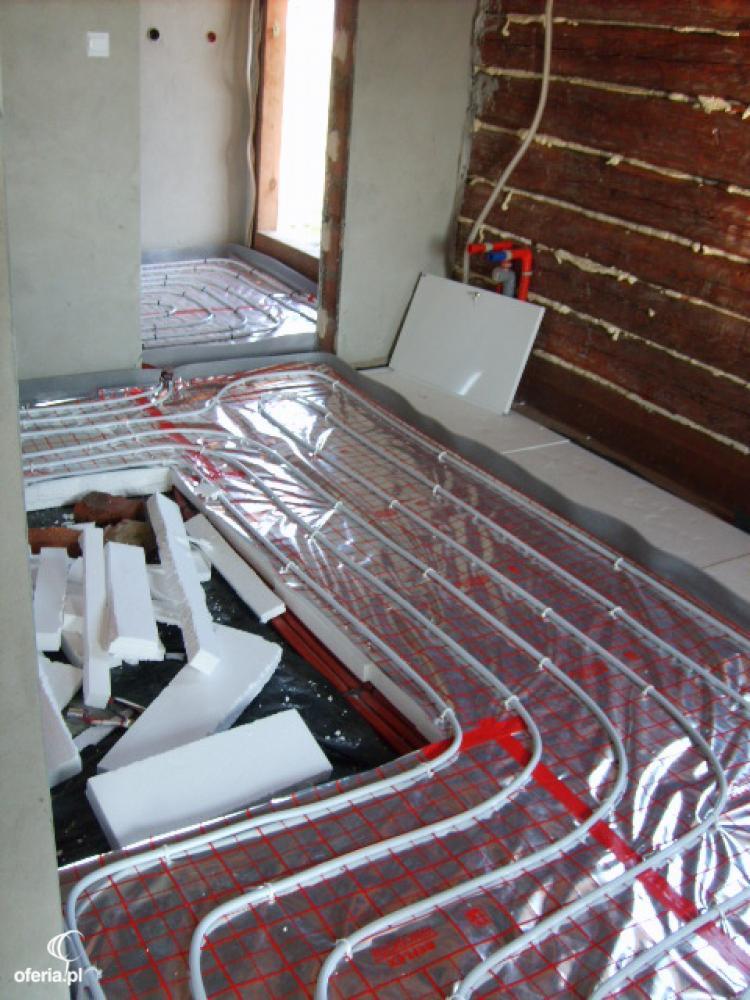 Montaż ogrzewania podłogowego: rury grzewcze rozłożone na folii z czerwoną siatką, fragment styropianu izolacyjnego, surowe ściany w trakcie wykończenia.