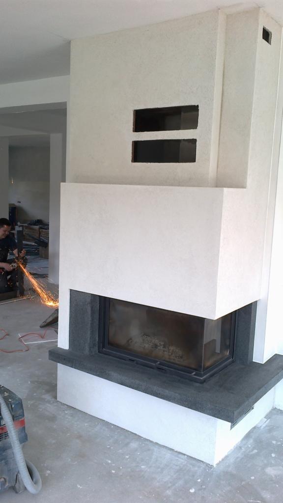Wnętrze w trakcie wykańczania z nowoczesnym kominkiem obudowanym białym tynkiem strukturalnym i ciemnym kamieniem, w tle pracownik używa szlifierki kątowej z iskrami.
