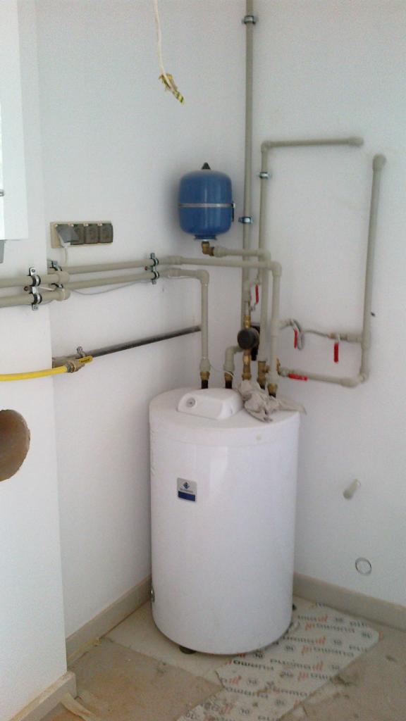 Biały, cylindryczny bojler z niebieskim zbiornikiem wyrównawczym i szarymi rurami w rogu białego pomieszczenia, widoczne przyłącza gazowe i elektryczne, podłoga zabezpieczona kartonem.