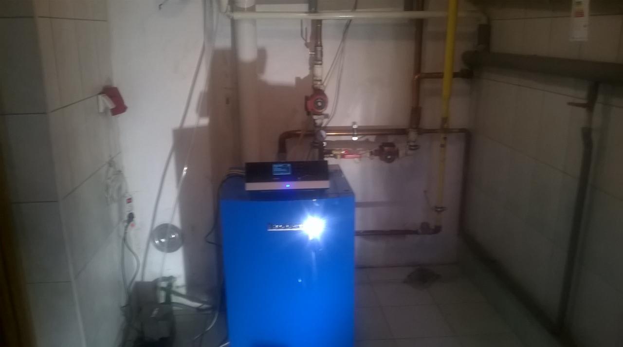 Niebieski piec gazowy z wyświetlaczem, zamontowany w pomieszczeniu z białymi płytkami, widoczne rury instalacji grzewczej i elementy sterujące.