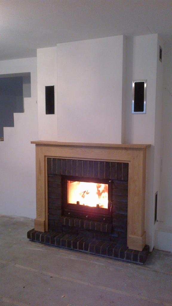 Nowoczesny kominek z cegły klinkierowej i drewnianą obudową, płomienie widoczne przez szybę, białe ściany z minimalistycznymi otworami wentylacyjnymi, surowa posadzka.