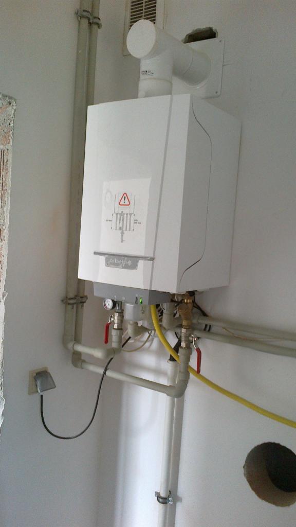 Biały, wiszący piec gazowy z podłączonymi rurami i kablem zasilającym, zamontowany na białej ścianie w trakcie instalacji, widoczny otwór w ścianie obok rur.