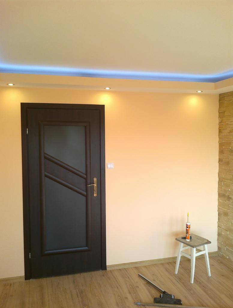 Pomarańczowe ściany w pokoju po odświeżeniu, widoczna czarna, przeszklona stolarka drzwiowa z ozdobnymi listwami, dekoracyjny kamień na ścianie, biały taboret z tubą silikonu i listwa przypodłogowa.