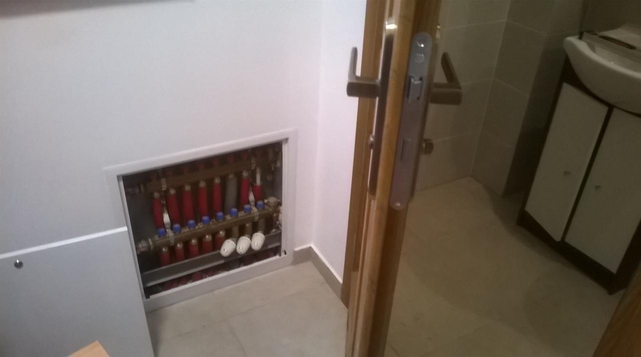 Rozdzielacz ogrzewania podłogowego ukryty za klapką rewizyjną w łazience, widoczne rury zasilające i powrotne, oraz zawory regulacyjne.