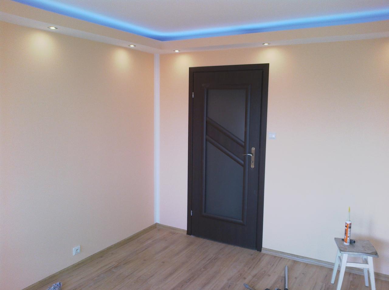 Pomalowane na beżowo ściany pokoju z nową, ciemną stolarką drzwiową i podwieszanym sufitem z niebieskim oświetleniem LED, widoczne ślady po pracach wykończeniowych.