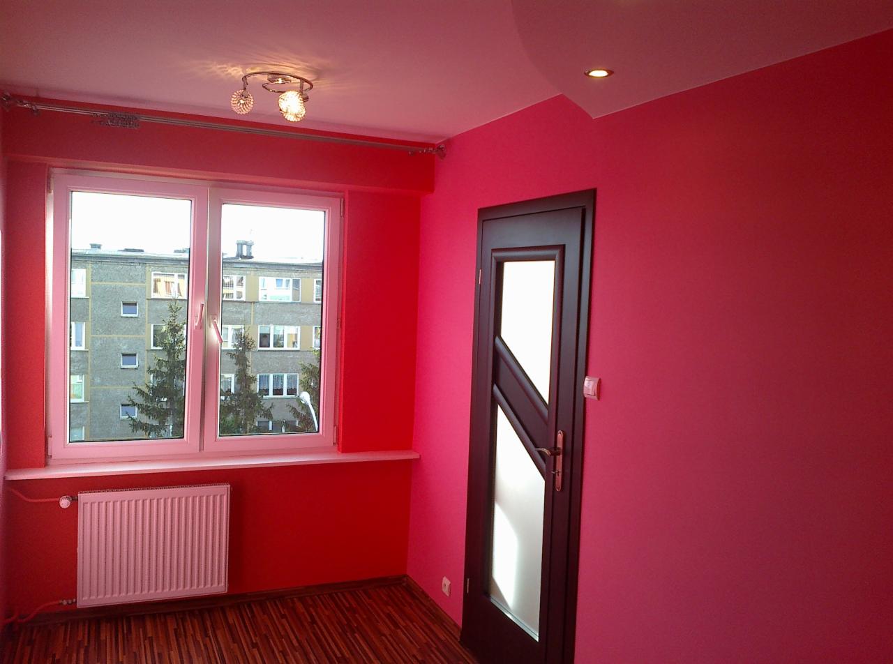 Pomieszczenie po malowaniu ścian na intensywny różowy kolor, widoczne okno z grzejnikiem, ciemne drzwi z przeszkleniem i drewniana podłoga.