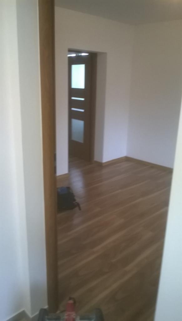 Pomalowane na biało ściany i nowa drewniana podłoga w pokoju, widoczne przez futrynę drzwi. Częściowo widoczny w rogu pokoju wózek malarski.