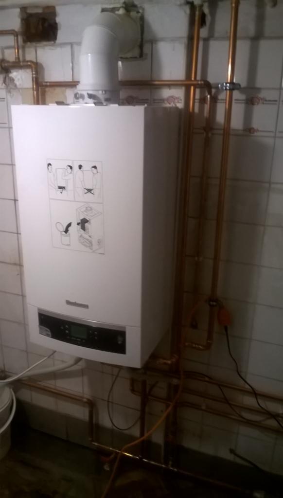 Biały piec gazowy Viessmann zamontowany na ścianie z białych płytek, widoczne miedziane rury instalacji, instrukcja obsługi na obudowie.