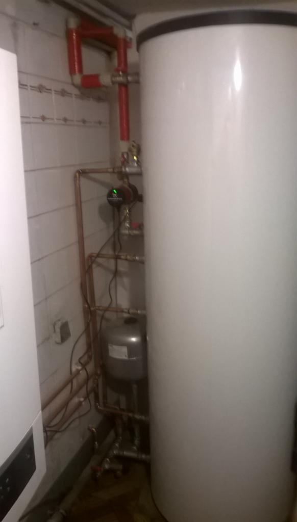 Bojler, zbiornik wyrównawczy i rury miedziane w kotłowni, widoczne połączenia z tworzywa sztucznego w kolorze czerwonym.